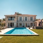 Wide angle shot of a 5-bedroom holiday house in countryside, large lawn and a swimming pool attached to the house. Blue sky and summer light.